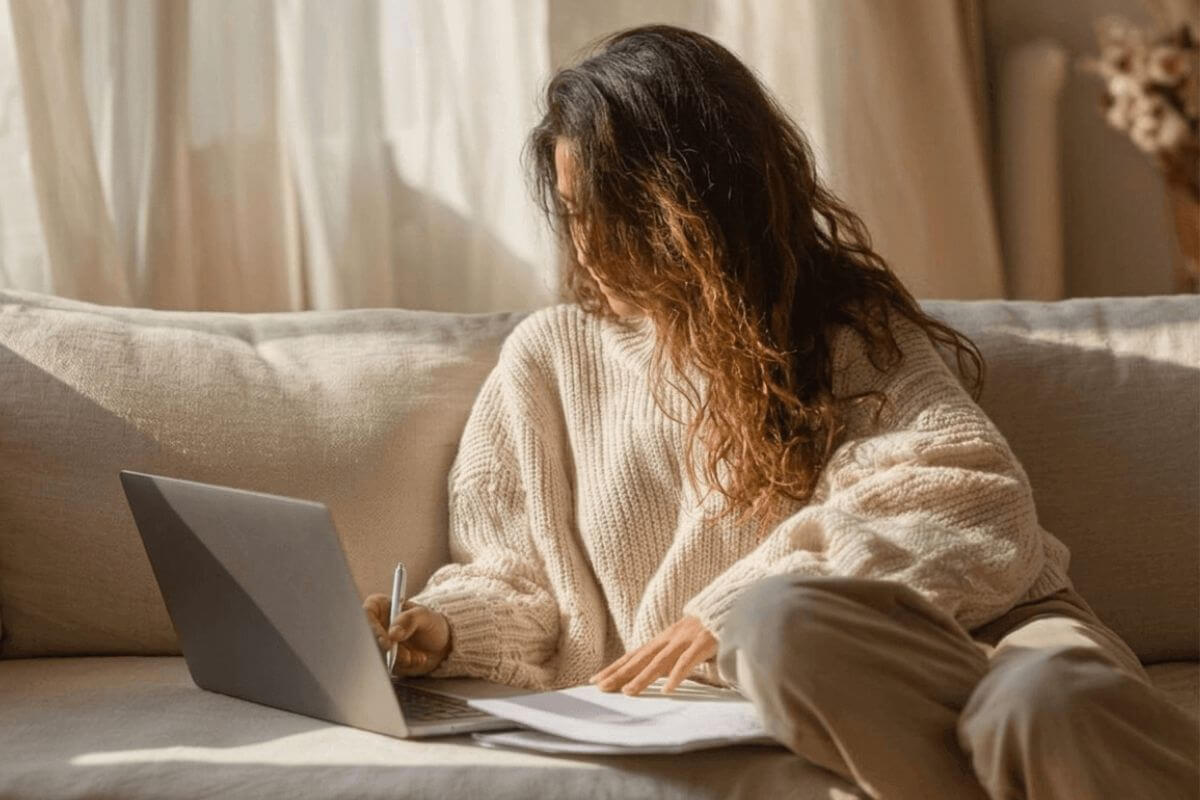 Young first-gen latina woman in a cream sweater sitting on a sofa with a laptop and papers, reviewing her finances and learning how to save money in your 20s
